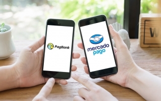 Mãos segurando celulares e mostrando na tela logos do PagBank e Mercado Pago